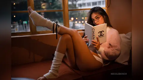 Young woman in pink sweater and white socks reading a Chanel book by a window in a cozy, warm indoor setting.