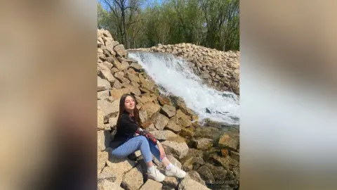 Smiling young woman in a black long-sleeve shirt and blue jeans sitting by a rocky waterfall in nature.