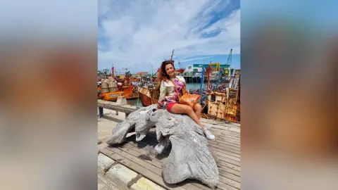 Smiling woman in a floral blouse and red shorts sitting on a stone sculpture at a sunny fishing harbor.