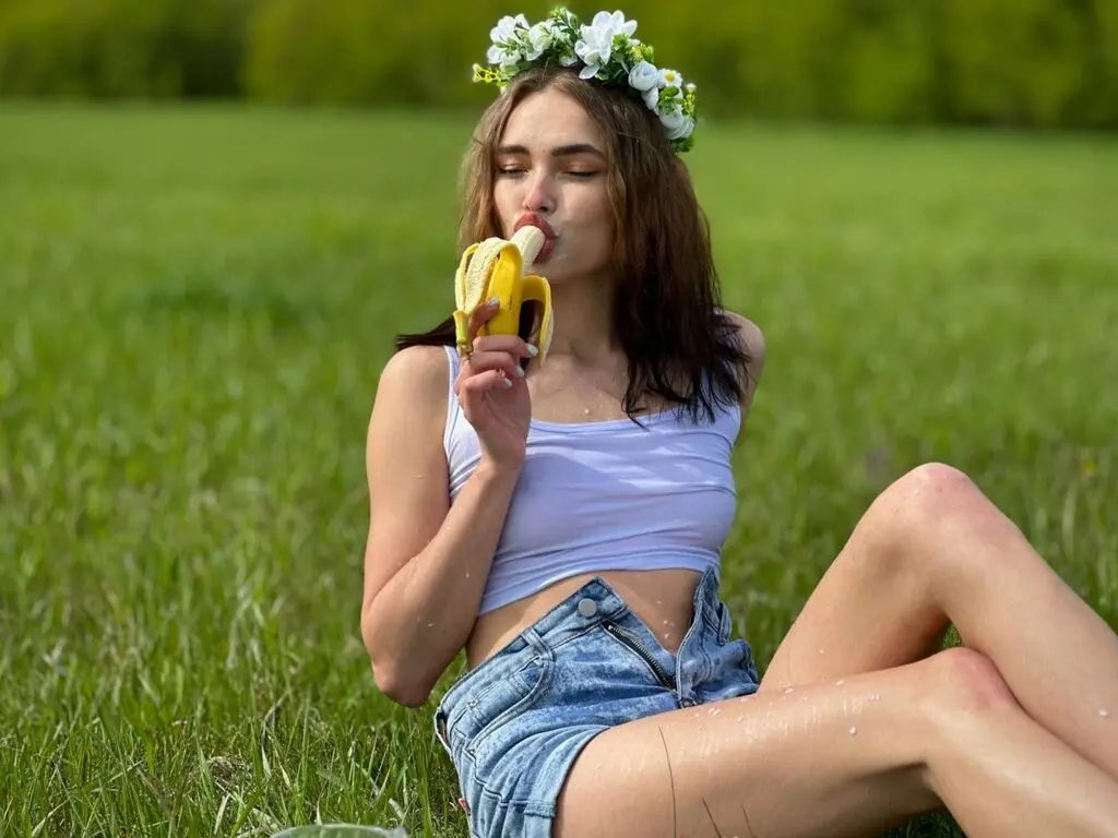 Young woman in white crop top and denim shorts eating a banana in a sunny green meadow.