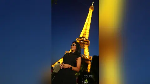 Dolce wears a black sleeveless dress and sunglasses while standing in front of the illuminated Eiffel Tower at night.