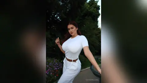 Elegant woman in a white t-shirt and trousers posing confidently in a lush green garden setting.