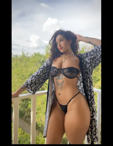 Curvy woman in a black floral kimono and lace lingerie posing on a sunny balcony overlooking lush greenery.