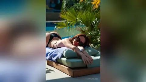 StacyHarland — Smiling woman in black bikini relaxing on a lounge chair by a sunny tropical resort pool.