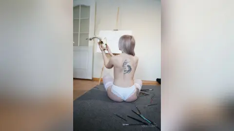 Blonde woman with a back tattoo sitting on a studio floor near an easel and art supplies.
