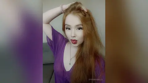 EpifaniaRalls has long red hair and wears a purple v-neck top while sticking her tongue out at the camera.