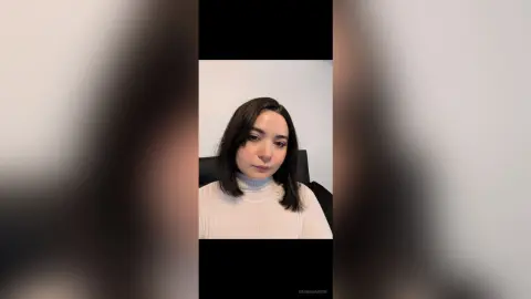 Young woman with dark hair wearing a white turtleneck sweater in a minimalist indoor setting.