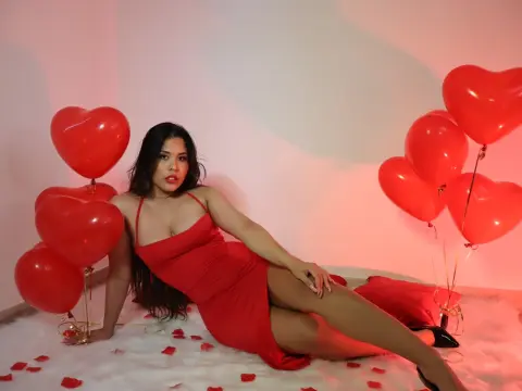 Woman in a red dress sitting on white fluff surrounded by red heart balloons in a romantic setting.