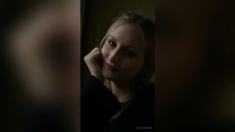 Moody portrait of a blonde woman in a black outfit looking thoughtfully into the camera in dim lighting.