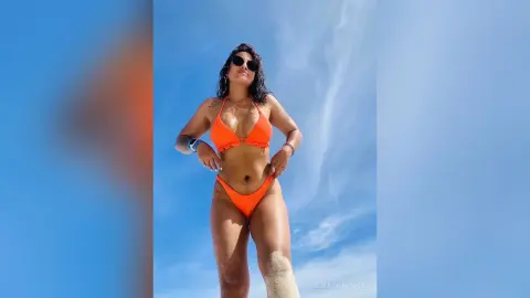 JulianaNavarro poses in an orange bikini against a clear blue sky, showing off her tanned skin and dark hair.