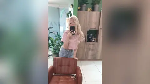 Blonde woman in a pink blouse and denim shorts taking a mirror selfie in a modern bright living room.