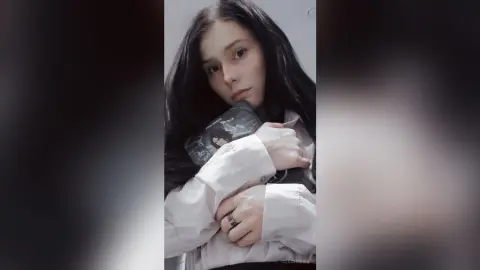 Young woman with long dark hair wearing a white long-sleeve shirt in a moody, minimalist indoor setting.