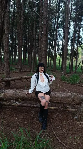 Woman in white ruffled blouse and black shorts holding a wine glass in a moody pine forest.