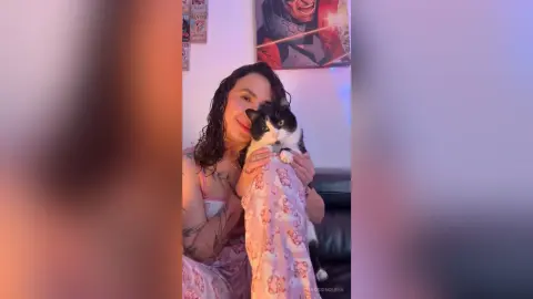 RossiNoleen smiles while holding a black and white cat close to her face in a pink patterned tank top.