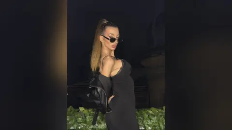 DeviNessa poses in a black dress and sunglasses, looking over her shoulder while holding a black leather jacket.
