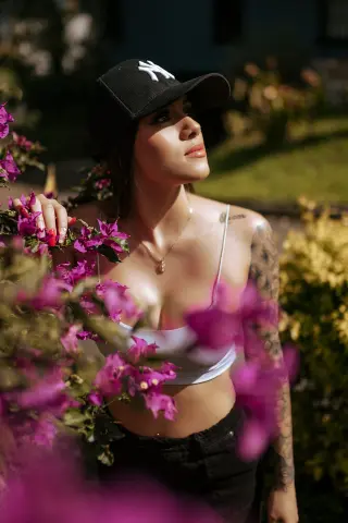 Young woman in a white tank top and black cap posing amidst vibrant pink flowers in a sunny garden.