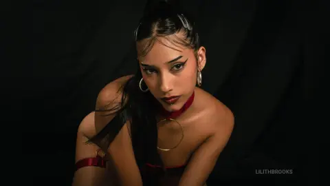 Young woman with winged eyeliner and hoop earrings posing in dark moody lighting wearing red lingerie.
