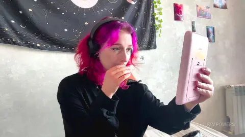 Young woman with pink hair wearing headphones looking into a mirror in a cozy bedroom setting.