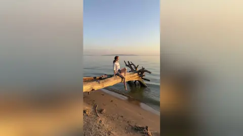 Woman in white top sitting on driftwood at a serene beach during a golden sunset.