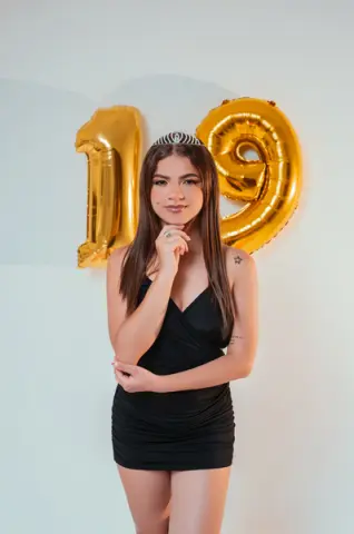 Young woman in a black cocktail dress celebrating her 19th birthday with gold foil balloons.