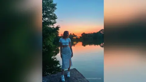 Young woman in a white crop top and lace skirt standing by a calm lake during a golden sunset.