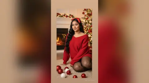 Smiling woman in a red sweater and headband sitting by a festive Christmas tree in a cozy holiday setting.