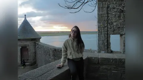 JessieLiu wears sunglasses and a tan jacket while leaning against a stone wall overlooking a body of water at sunset.