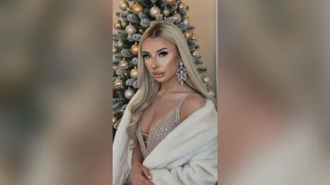 Glamorous blonde woman in a sparkling sequin dress and white faux fur coat posing by a festive Christmas tree.