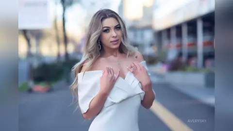 Blonde woman in a white off-the-shoulder dress posing on a city street with a soft, dreamy mood.