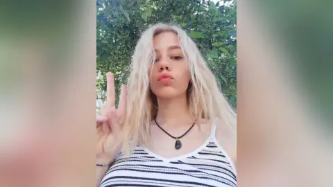 Blonde woman in a striped tank top making a peace sign outdoors against a lush green leafy background.