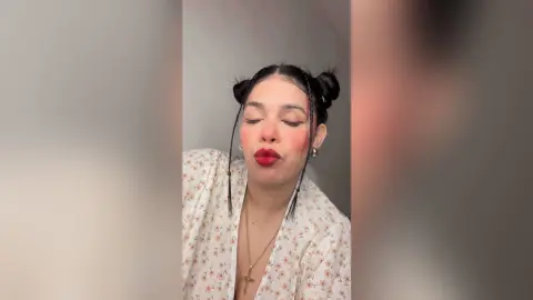 Young woman with braided buns and red lipstick wearing a floral blouse in a soft, minimalist indoor setting.