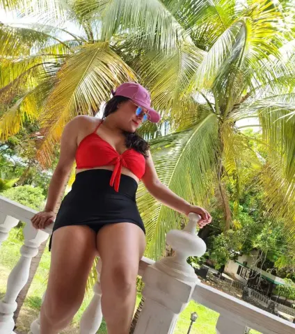 Woman in red bikini top and black high-waisted bottoms posing by palm trees on a sunny tropical balcony.