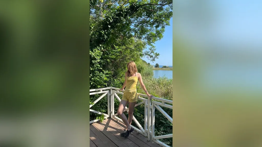 MozelleEzzo wears a blue floral sundress while posing outdoors by the water on a sunny day.