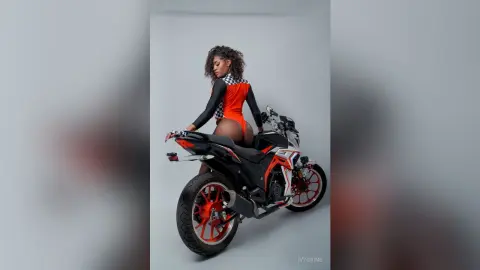 Stylish woman in a red and black bodysuit posing confidently on a modern black motorcycle against a grey backdrop.