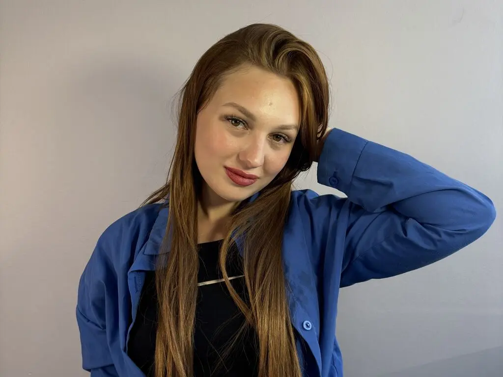 DorisDonachie poses with her long brown hair tucked behind her ear, wearing a blue button-down shirt over a black top against a plain grey background.