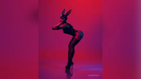 Woman in black bunny mask and bodysuit posing against a moody red and purple studio background.