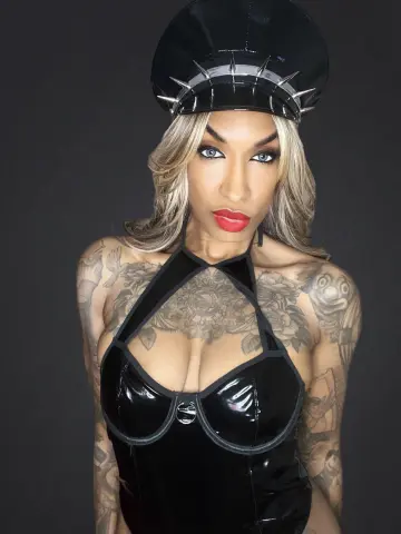 Tattooed woman in black latex bodysuit and spiked cap posing against a dark background.