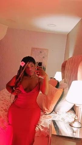 ManelykStorm poses in a red satin dress with a pink flower in her long dark braids while sitting on a bed.