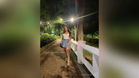 Young woman in a white corset top and blue denim skirt walking on a park path at night.