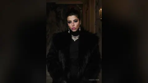 Elegant woman in a black faux fur coat and statement necklace posing in a moody, dimly lit luxury setting.