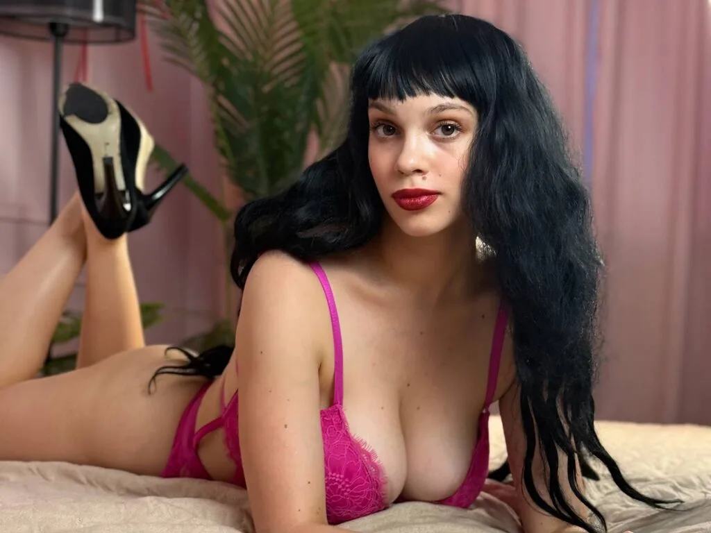 ErikaBradley lies on a bed wearing pink lace lingerie and black high heels.