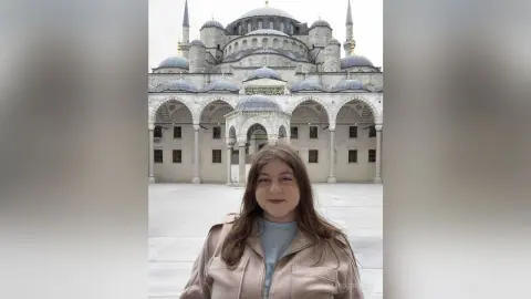 Smiling woman in a beige jacket standing before the historic Blue Mosque in Istanbul on a bright day.