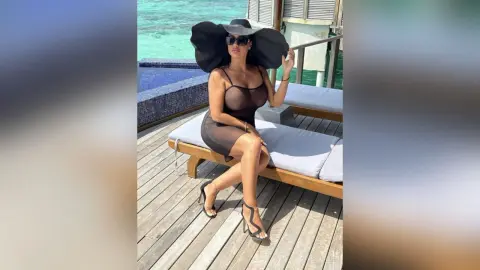 MissyJolie sits on a white lounge chair wearing a tight brown sleeveless dress and a large black sun hat.