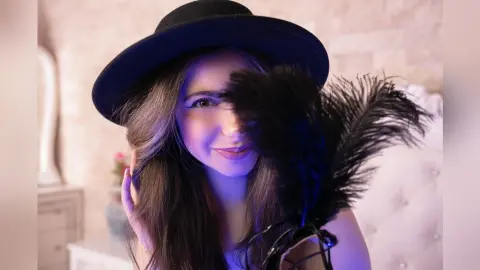Smiling woman in a black hat and feathered accessory posing in a soft, dreamy indoor setting.