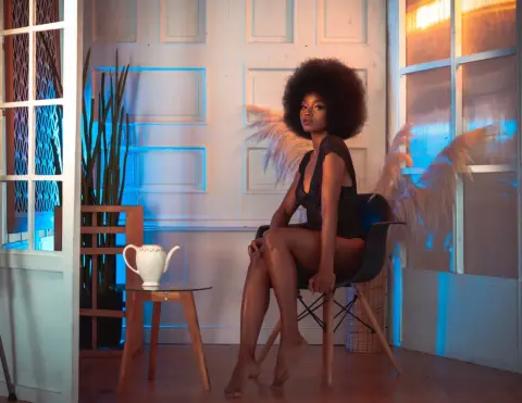 Elegant woman in a black dress with feathered wings sitting in a moody, blue-lit room by a white tea set.