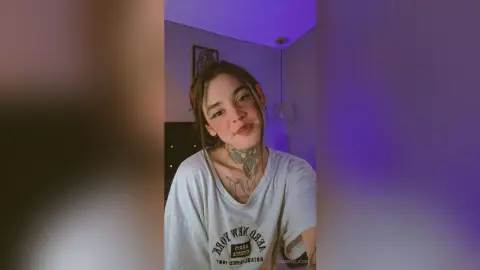 Smiling young woman with neck tattoos wearing a white graphic t-shirt in a dimly lit room with purple lighting.