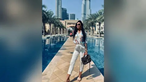 Stylish woman in white trousers and patterned blouse posing by a luxury city pool with skyscrapers in the background.