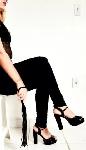 Elegant woman in black lace bodysuit and trousers sitting in a bright minimalist room wearing high heels.