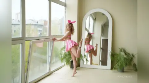 Cheerful woman in pink lingerie posing playfully by a bright window in a sunlit modern room.