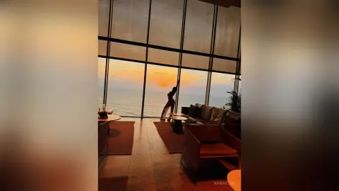 Silhouette of a woman standing by a large window overlooking a sunset ocean view in a modern luxury living room.